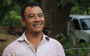 Un hombre sonriente se encuentra de pie frente a una valla de madera, mostrando un semblante alegre en un entorno al aire libre.