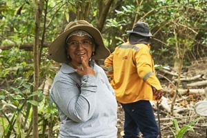 Una mujer con sombrero está de pie junto a un hombre en el bosque, rodeados de árboles y vegetación natural.