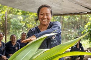 Una mujer sonriente con una camisa azul manipula grandes hojas verdes en un entorno soleado al aire libre, rodeada de árboles y personas sentadas al fondo.