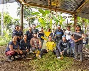 Un grupo de personas posa alegremente bajo un refugio rústico, rodeado de exuberante vegetación y plataneros.