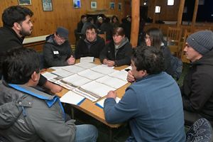 Un grupo de ocho personas se sienta alrededor de una mesa de madera en una habitación con iluminación cálida, inmersas en una seria conversación. Papeles y folletos cubren la mesa.