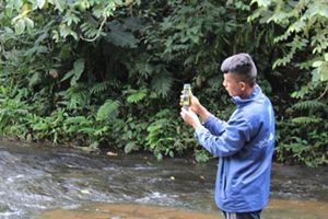 Una persona con chaqueta azul está de pie junto a un arroyo, examinando un dispositivo portátil. 