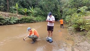 Tres personas realizan trabajo de campo en un río poco profundo y de color marrón, rodeado de exuberante vegetación. Una toma muestras de agua, otra toma notas y la tercera observa el agua.
