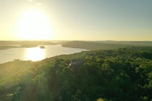  Vista aérea de un río iluminado por el sol que serpentea a través de un denso bosque verde al atardecer, con reflejos dorados y un paisaje tranquilo y extenso.