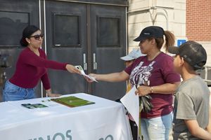 A person behind a table, wearing a cover with "Latino Progresando" on it, hands a pamphlet to another woman who is there with a child. 