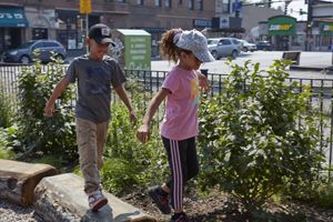 Two children walk on flat, square stones among a garden in a schoolyard. 