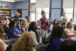 A woman standing, speaks through a microphone while other people are seated around her. 