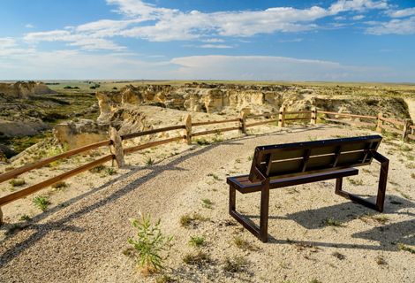 A wood bench sits at an overlook point at Little Jerusalem Badlands State Park on a clear day. In the distance is a view of rock formations and prairie.