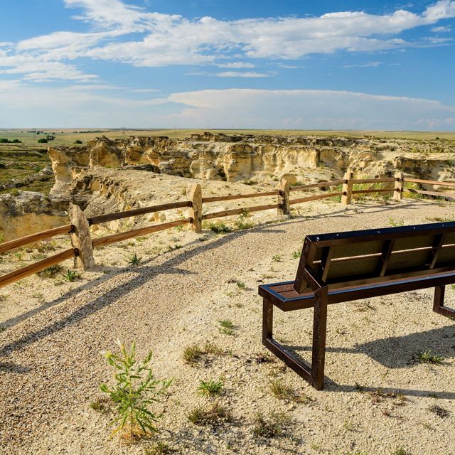 A wood bench sits at an overlook point at Little Jerusalem Badlands State Park on a clear day. In the distance is a view of rock formations and prairie.