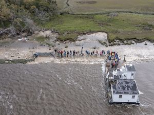 Several people stand along a shoreline in front of a white barge.