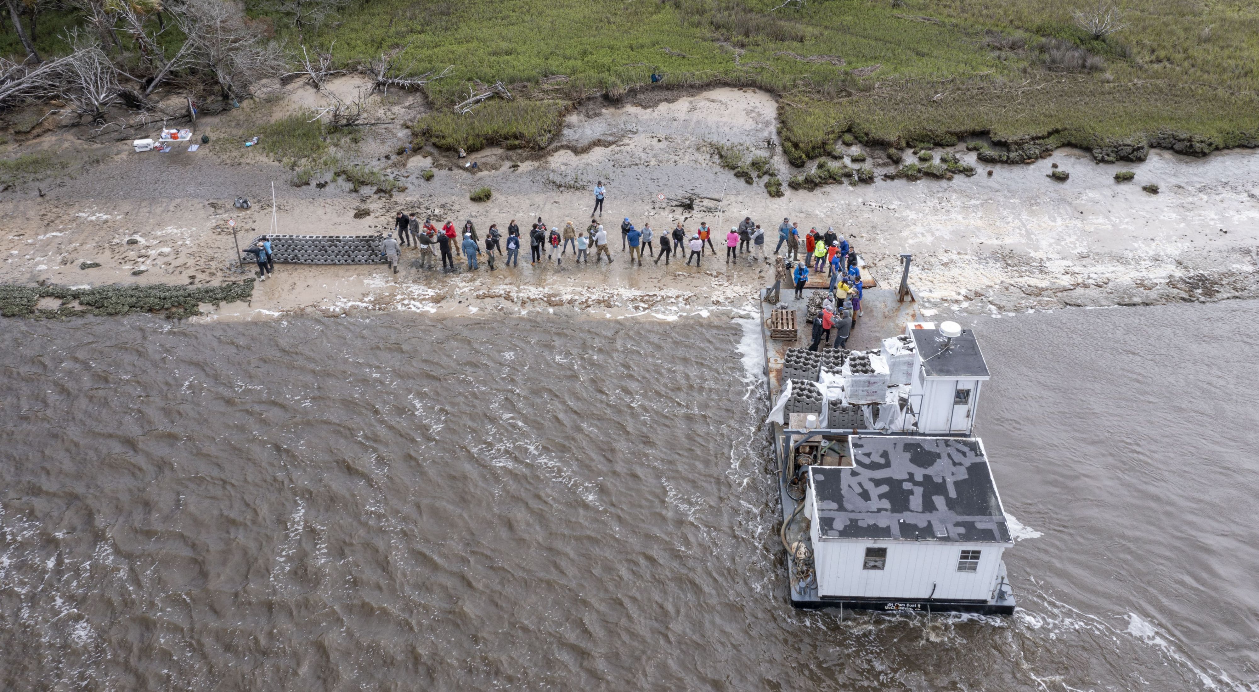 An aerial view of several people standing along a shoreline in front of a white barge.