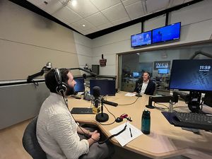 Two people sit across a table from each other in conversation in a recording studio, with mics and headphones.