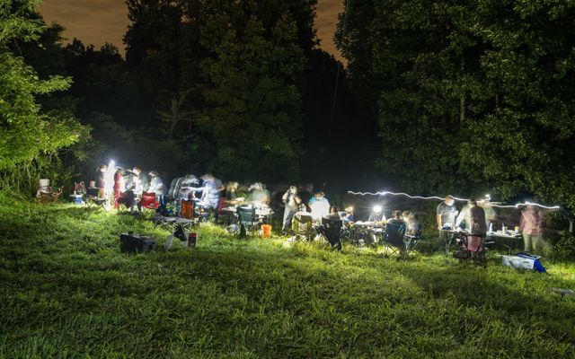 A group of more than a dozen people stand around tables in a green grass area at nighttime, their flashlights and headlamps illuminating their workstations.