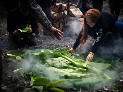 A person with long red hair assembles long green leaves over a smoking section of ground.