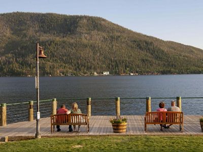 Two pairs of people sit on park benches overlooking Grand Lake, Colorado, with a tree-covered mountain in the background.