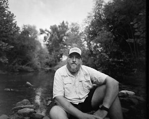 Black-and-white image of Jason Throneberry sitting at the edge of a river.