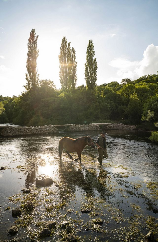 A man walks a horse in a river shallows.