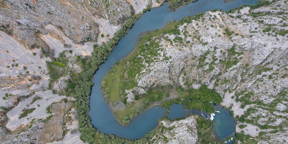 Aerial view of a winding river lined with green bushes and surrounded by rocky areas.