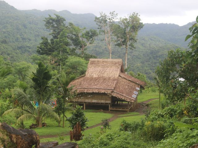 A typical house in a village in the Solomon Islands, with a roof made from Sago palm tree leaves and surrounded by lush forest and mountains.