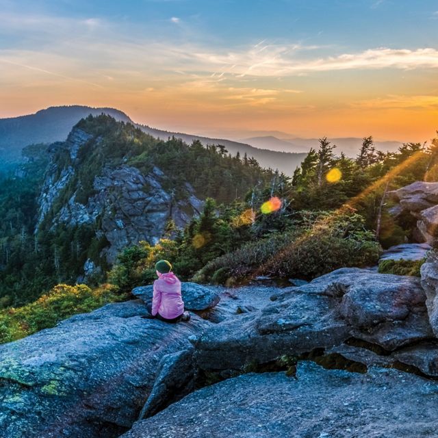 Frances experiences sunrise at Attic Window after camping on Grandfather Mountain ridge.

Ethan,
Thanks, I’m honored to be included in the running.
Here’s the info you requested:
Jim Magruder
P.O. Box 154, Linville, NC 28646

Photo Caption:
Grandfather Mountain Sunrise
The photographer’s wife Frances takes in sunrise from the Attic Window peak on Grandfather Mountain, North Carolina. 
Through the combined efforts of The Nature Conservancy, the state of North Carolina, and the owners of Grandfather Mountain, Inc. Grandfather Mountain State Park was established in 2009 to preserve and protect Grandfather, one of the highest peaks in the Blue Ridge Mountains and one of the oldest mountains in North America. Grandfather Mountain is home to a wide variety of rare plants and animals and offers some of the most rugged and spectacular hiking in the eastern US.

This image is a two exposure HDR merge. One exposure for the sunrise sky and one for the rocky foreground. I cropped the merged HDR image to a panorama format to eliminate  some bland sky and rock at top and bottom.

Because of email size restrictions I am sending each of the 2 RAW files and the hi-res JPEG as attachments to separate emails.

Thanks,
Jim