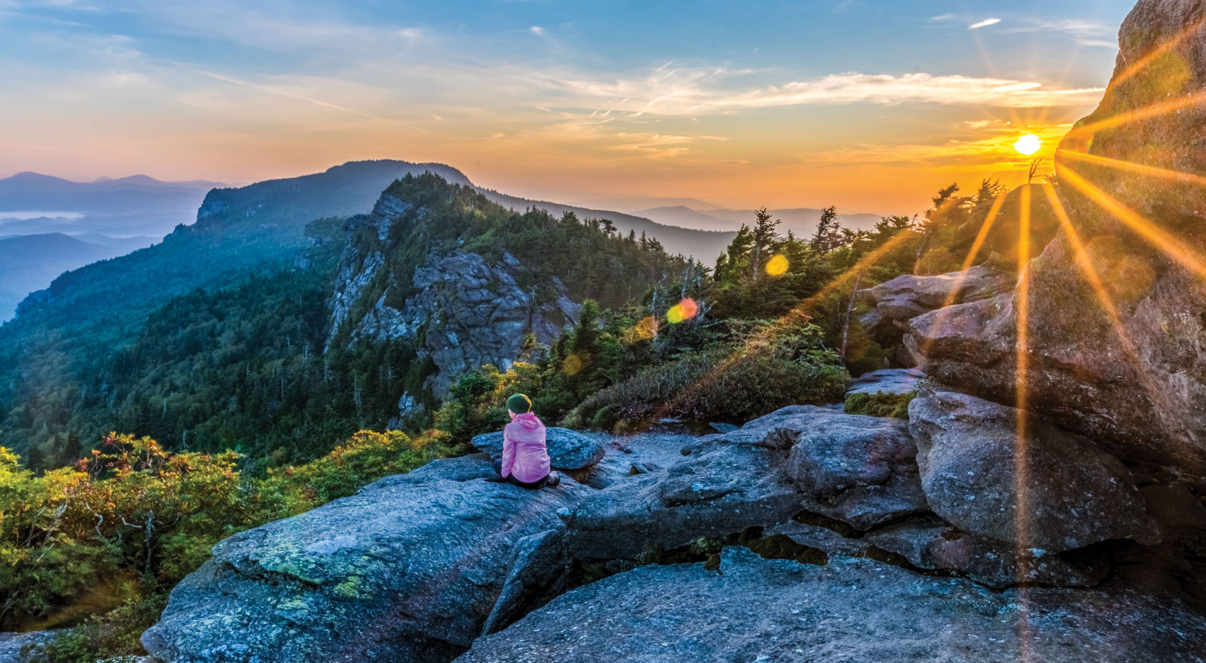 Frances experiences sunrise at Attic Window after camping on Grandfather Mountain ridge.

Ethan,
Thanks, I’m honored to be included in the running.
Here’s the info you requested:
Jim Magruder
P.O. Box 154, Linville, NC 28646

Photo Caption:
Grandfather Mountain Sunrise
The photographer’s wife Frances takes in sunrise from the Attic Window peak on Grandfather Mountain, North Carolina. 
Through the combined efforts of The Nature Conservancy, the state of North Carolina, and the owners of Grandfather Mountain, Inc. Grandfather Mountain State Park was established in 2009 to preserve and protect Grandfather, one of the highest peaks in the Blue Ridge Mountains and one of the oldest mountains in North America. Grandfather Mountain is home to a wide variety of rare plants and animals and offers some of the most rugged and spectacular hiking in the eastern US.

This image is a two exposure HDR merge. One exposure for the sunrise sky and one for the rocky foreground. I cropped the merged HDR image to a panorama format to eliminate  some bland sky and rock at top and bottom.

Because of email size restrictions I am sending each of the 2 RAW files and the hi-res JPEG as attachments to separate emails.

Thanks,
Jim
