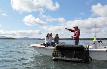 Three people sit at the bow of a small boat. A fourth person stands in the middle of the boat, pointing to something out of frame behind them.