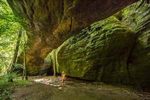 A person stands underneath a huge arch of rock that acts like a canopy amid a forest.
