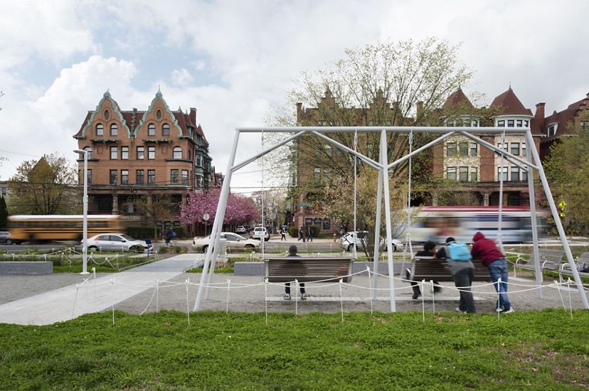 Three people sit on a swing set in a small grassy area on a city street.