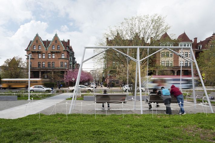 A group of people sit on a swing set in a small area of grass on a city block.