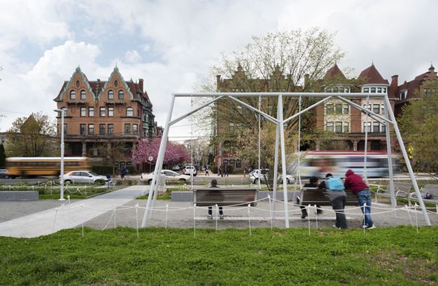 Three people sit on a swing set in a small grassy area on a city street.