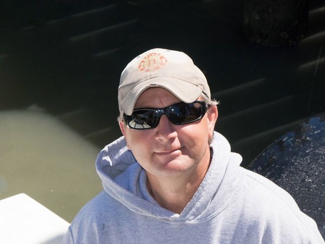 A man looking up at the camera from the deck of a small jonboat. He is wearing a gray hoodie and sunglasses. The sun reflects on the still water behind him.