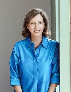 A woman with blue shirt leans against a white wall.