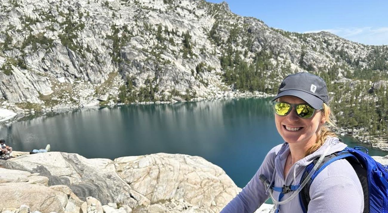 A smiling woman poses for a selfie in front of a blue mountain lake.