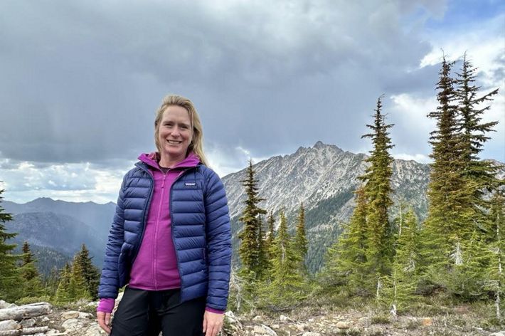A woman stands in a rocky clearing. A sharp mountain peak rises behind her.