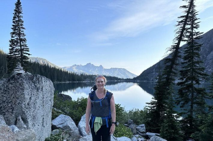 A woman stands in front of a mountain lake. The still surface of the water reflects the towering mountain peaks around it.
