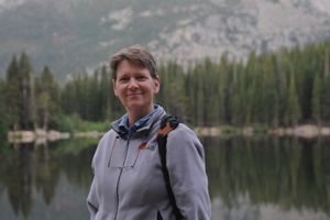 Mary Huffman standing on the shore of a lake with treeline and mountain in the background