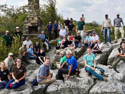 A group of two dozen people pose together during a Fire Learning Network conference outing. People sit and stand on a large rocky outcropping. 