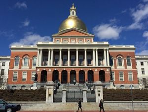 View of the Massachusetts State House with its gold dome from across the street on a blue sky sunny day.