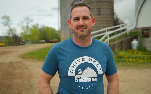 Matt Tentis standing in front of his family farm's barn wearing a t-shirt that reads White Barn Acres.