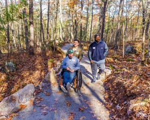 A man and his friend push another man using a wheelchair down a flat, winding path in the woods.