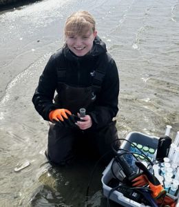 A young woman in a wet suit kneels in a shallow tidal creek next to an assortment of scientific equipment