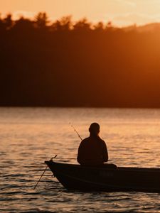 A silhouetted person sits with her back to the camera in a small boat holding a fishing rod in a body of water. 