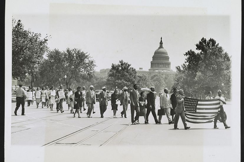 A group of Black Americans march together along an open plaza. Two men leading the group are in uniform and carry an American flag. The dome of the US Capitol building rises in the background.