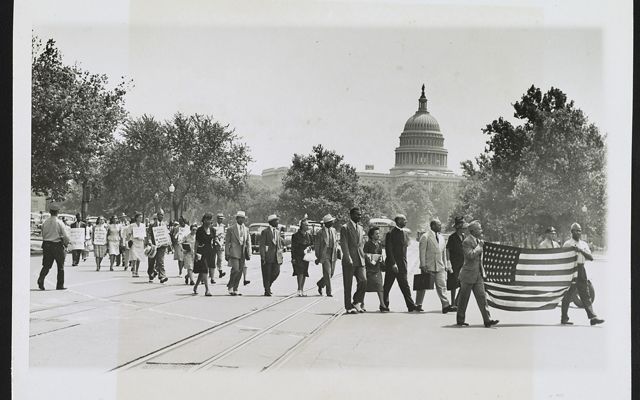 A group of Black Americans march together along an open plaza. Two men leading the group are in uniform and carry an American flag. The dome of the US Capitol building rises in the background.