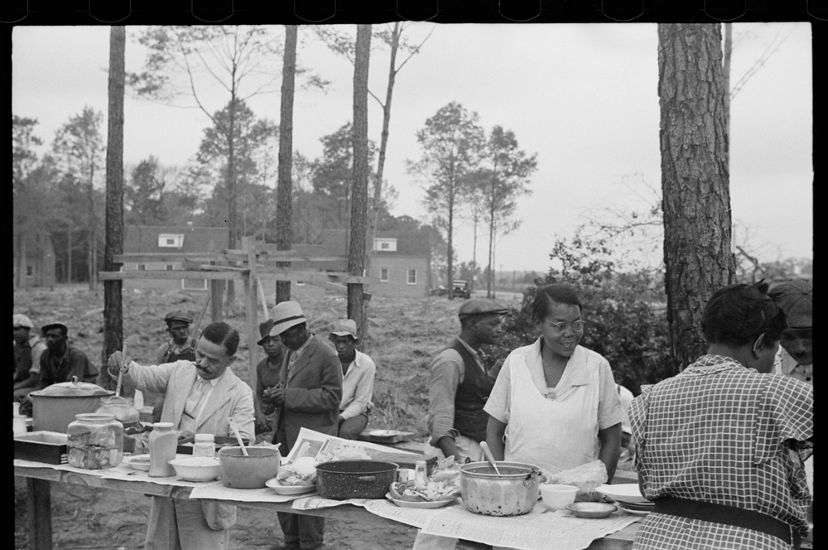 Black and white photo from the 1930s. A group of people enjoy an outdoor meal. Dishes are set up buffet style on rough cut boards. 