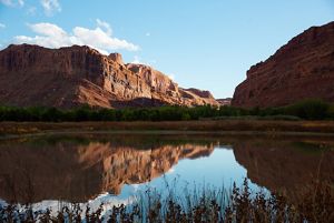 Two large red rock formations loom above a body of still water and cast reflections on the water's surface.