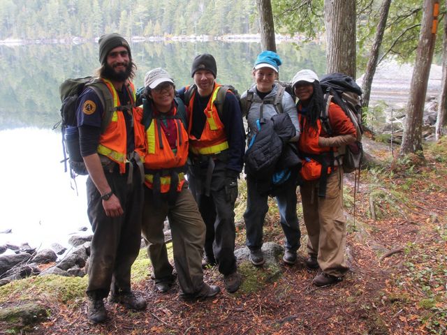 Five trail builders with backpacks pose together in front of a lake.