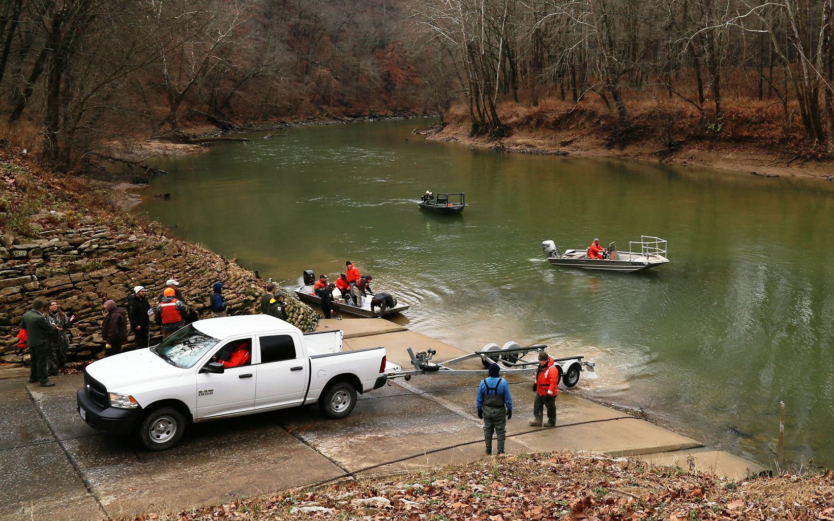 A white truck surrounded by people backs up to a river.