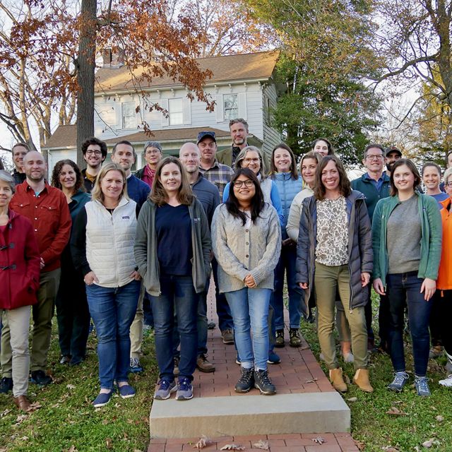 Group photo of TNC MD/DC staff. A large group of people stand together outdoors in front of a backdrop of trees, smiling during the chapter's annual staff retreat.