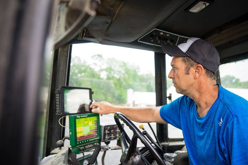 A man sits in the cab of a tractor. Two small computer display screens sit in front of him behind the steering wheel providing information to help him apply fertilizer to his crop.